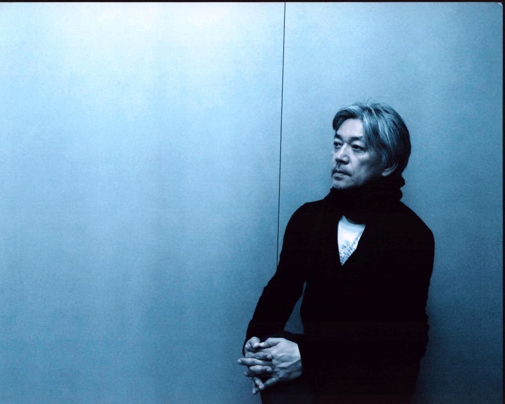 Ryuichi Sakamoto photo print in black sweater and scarf in blue room.