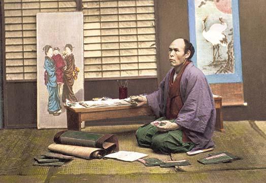 Japanese artist kneeling by bench with materials, hand-tinted Meiji era photograph.