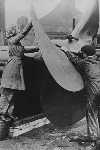 Poster print of English workers laboring over a ship's propeller at a shipyard.