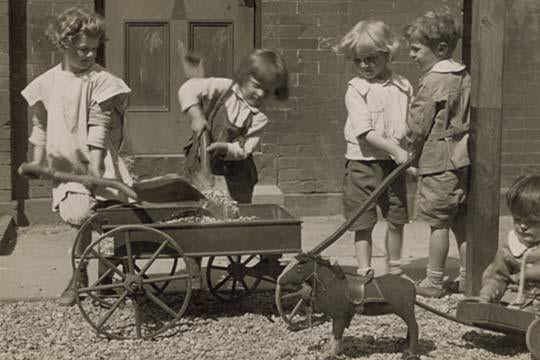 Poster print of four young children loading rocks into a wagon while a smaller child plays with a toy pony cart.