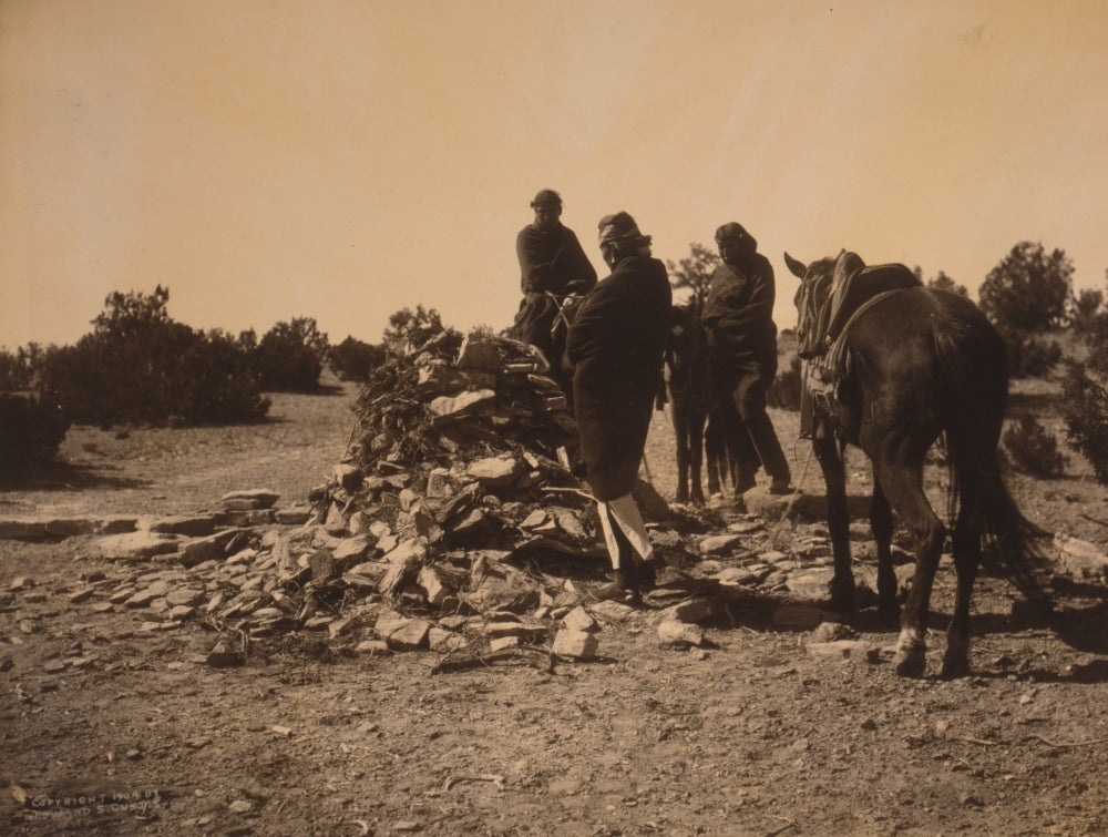 Three Navajos with horses gathered before a mound of rocks and vegetation.