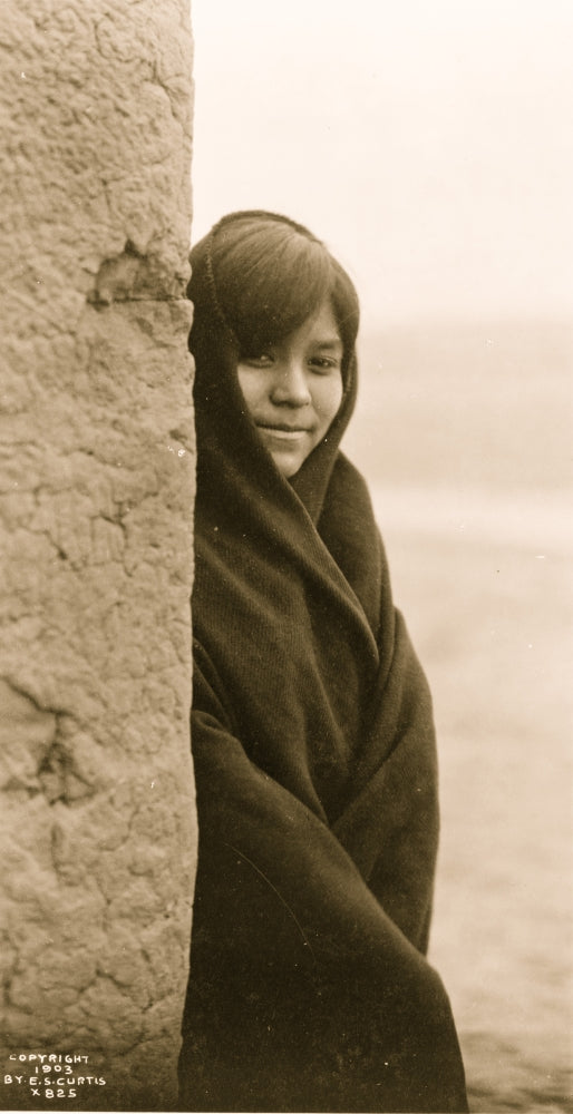 Three-quarter length portrait of a Zuni girl peering around a building.