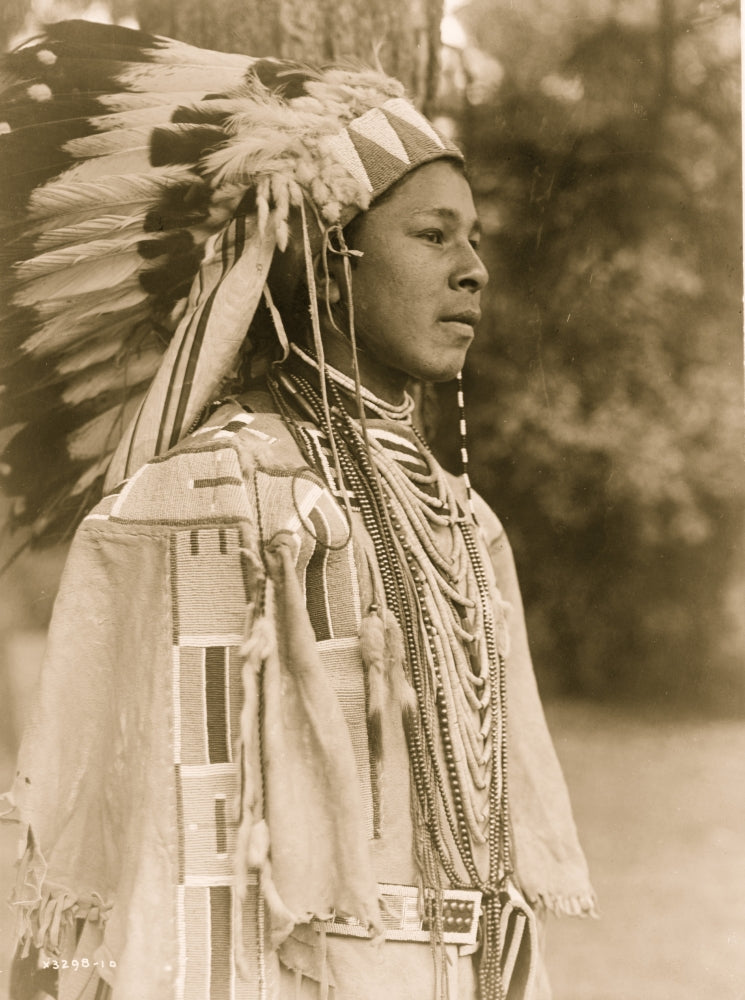 Half-length portrait of Umatilla youth in full feather headdress and beaded shirt.