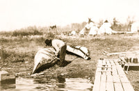 Ojibwa Indian mending canoe at shore with tepees in background print.