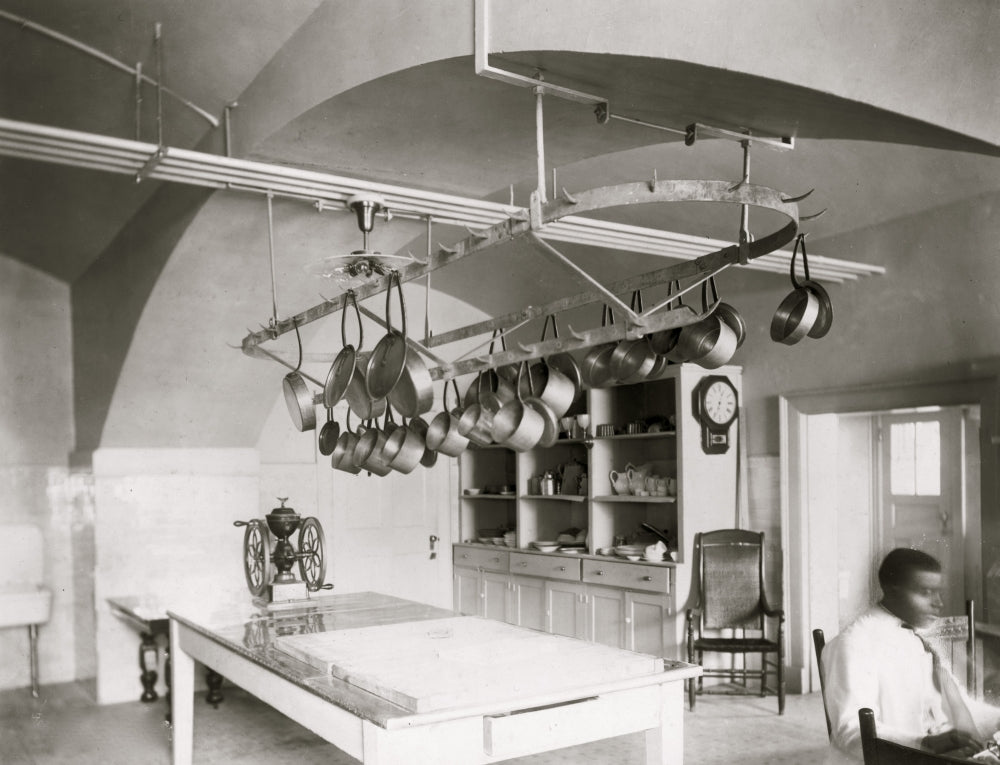 African American man seated at desk beside kitchen table with pots hanging.