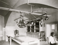 African American man seated at desk beside kitchen table with pots hanging.