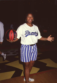 Queen Latifah holding a basketball, wearing blue striped shorts and a Brooklyn shirt.