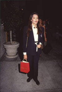 Barbara Carrera photo print in black pants and jacket holding a red shopping bag
