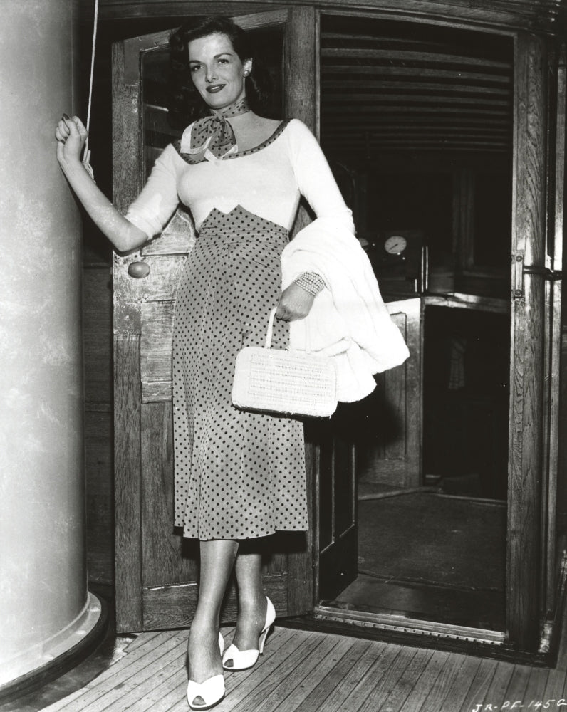 Jane Russell posed in a scoop-neck long sleeve dress with polka dot high waist skirt holding a white handbag.
