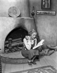 Bette Davis seated by the fireplace, holding a cigarette and reading a book.