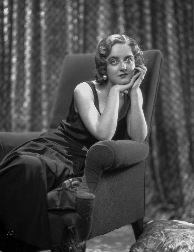 Bette Davis seated on a black couch in a black velvet dress, classic Hollywood photo print.