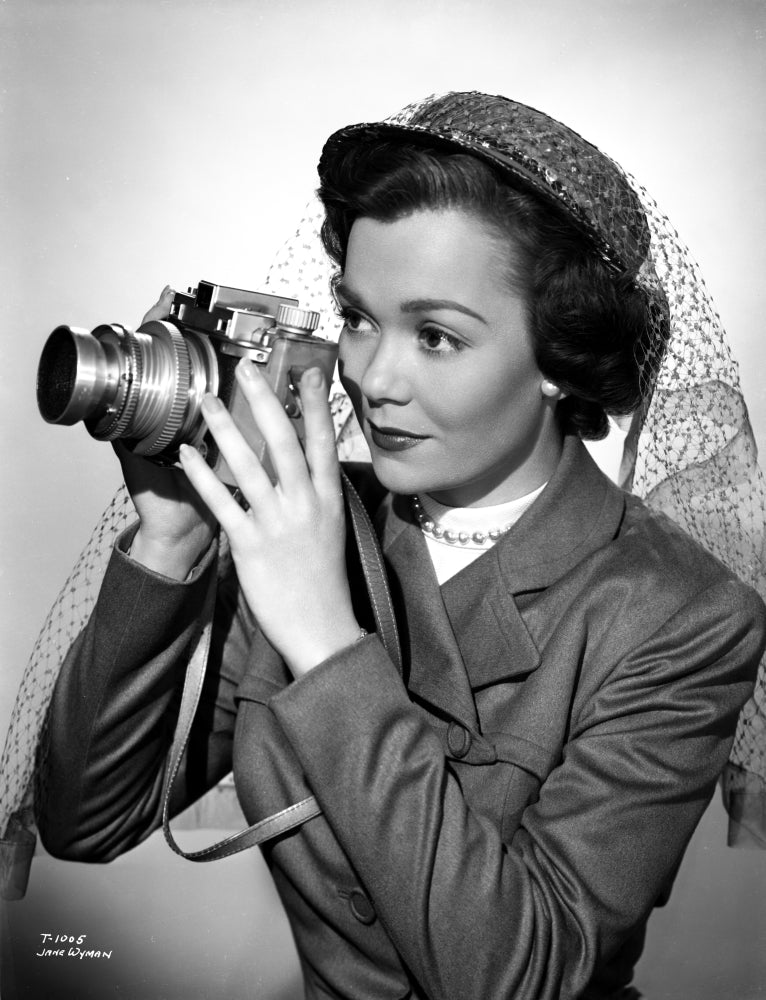 Portrait of Jane Wyman in grey linen suit holding a camera.