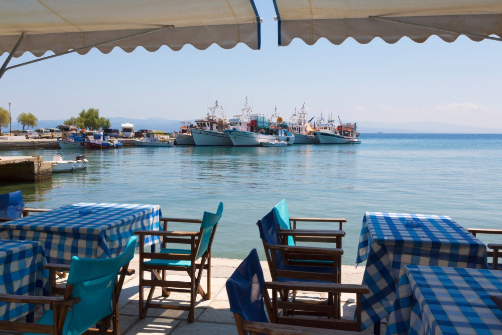 Poster print of Platanias, Thessaly, Greece showing a harbour scene with tables, chairs, and fishing boats.