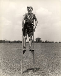 1930s-1940s print of a smiling boy on stilts wearing a striped shirt.