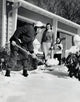 Father shoveling snow in front yard with family watching.