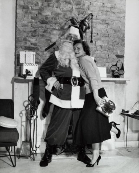 Poster print of a young woman holding a Christmas present and hugging Santa Claus in front of a fireplace.