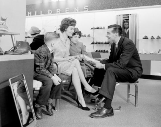 Shop assistant showing women's shoe to mother with son and daughter poster print.
