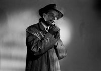 Portrait of a young man wearing a coat and hat, smoking a cigarette in a studio shot.