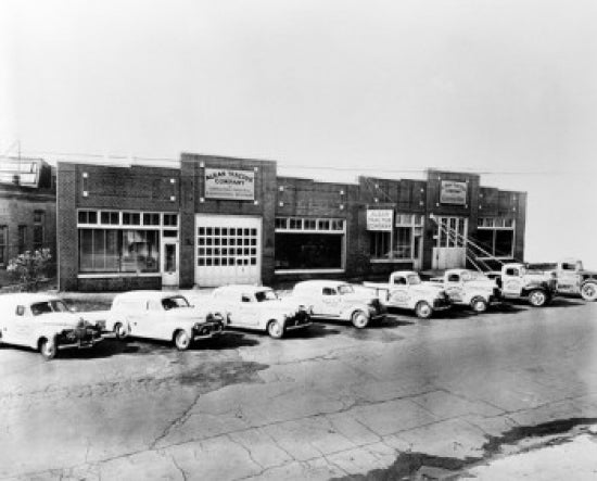 Trucks in front of Alban Tractor Co factory poster print, showcasing vintage industrial scene.