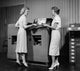Side profile of two businesswomen standing in front of a machine poster print.
