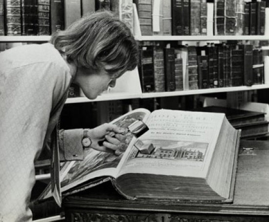 Young man examining a miniature Bible in a library setting poster print.
