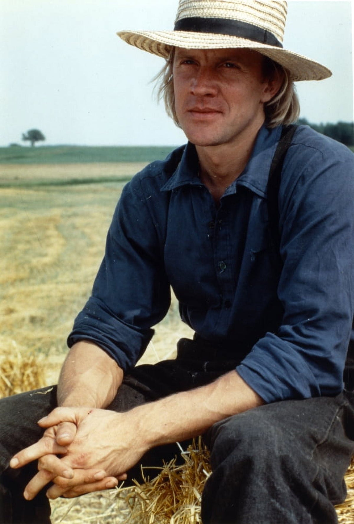 Alexander Godunov photo print sitting on hay stack in blue long sleeves and hat.